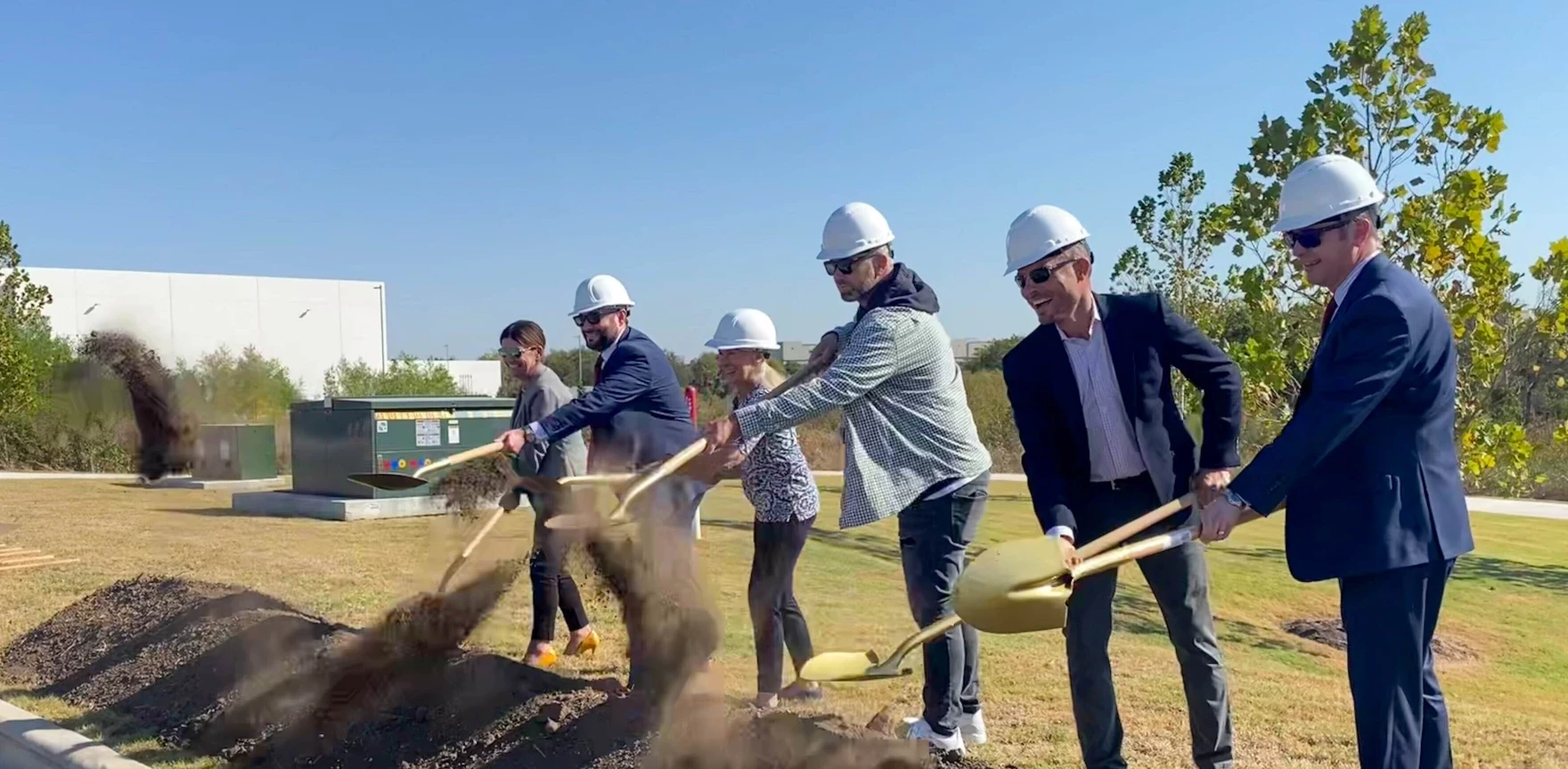 Six people in nice clothes and white hardhats turning dirt with golden shovels