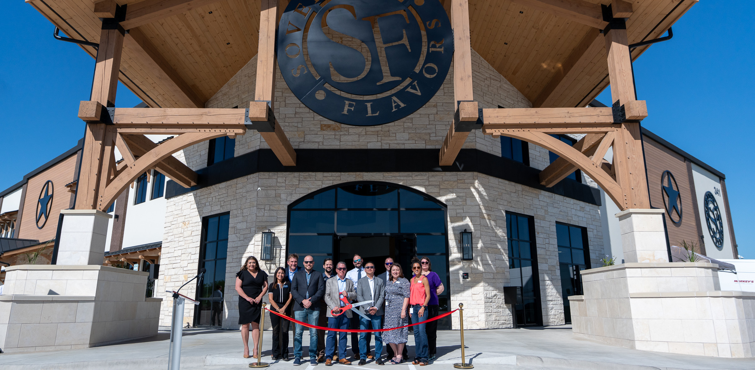 Sovereign Flavors and City of Kyle leadership and staff stand in front of new building with large scissors poised to cut red ribbon.