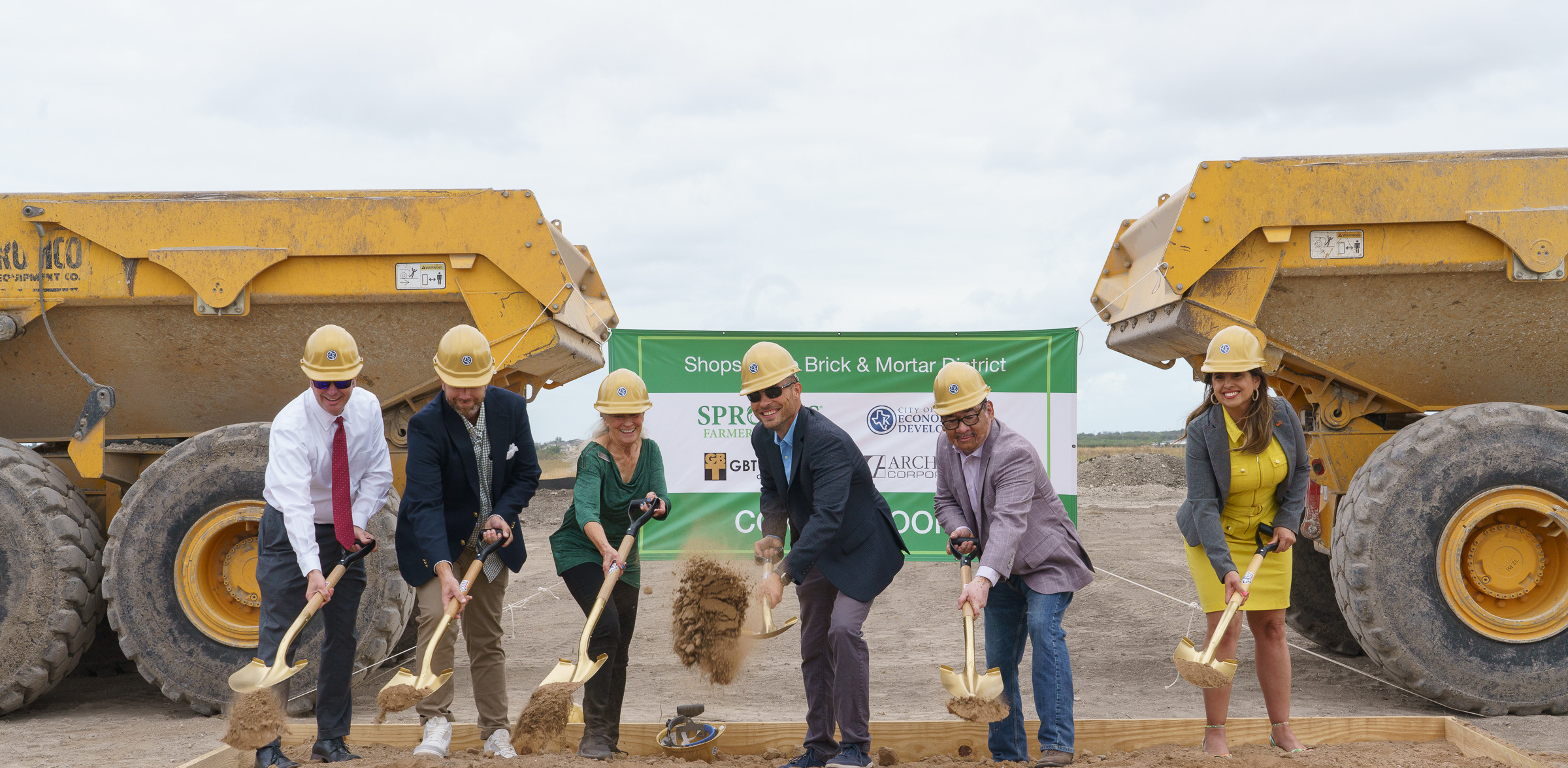 Four men and two women shoveling dirt with gold shovels in front of two construction vehicles and an obscured sign.