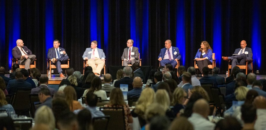 Panel of men and women in business attire sitting on chairs on a stage in front of a crowd