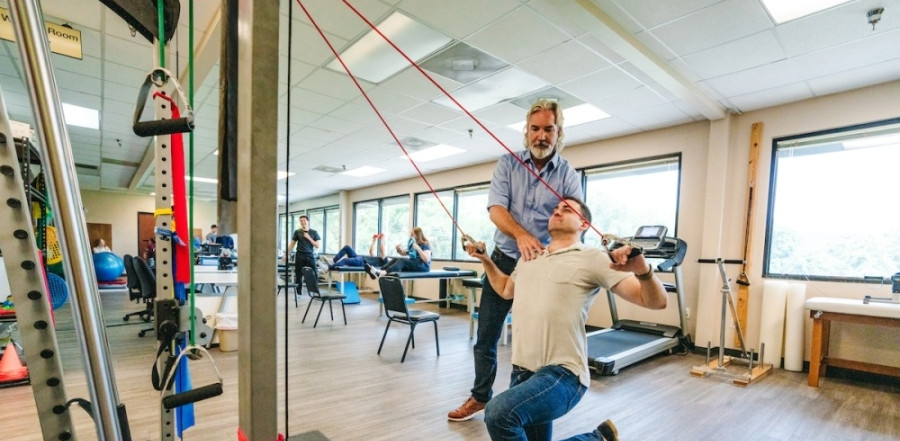 Man helping another attain correct position while doing an exercise. Other people and workout equipment in the background.